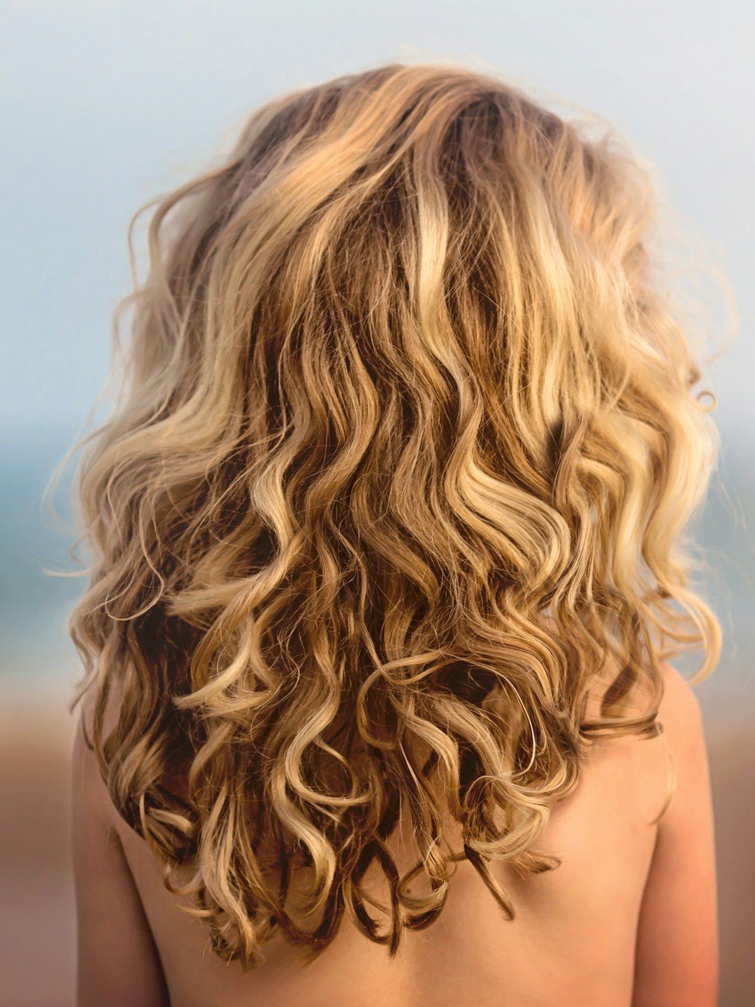 Cabelo loiro mel, longo e cacheado, visto de costas contra a luz do fim de tarde