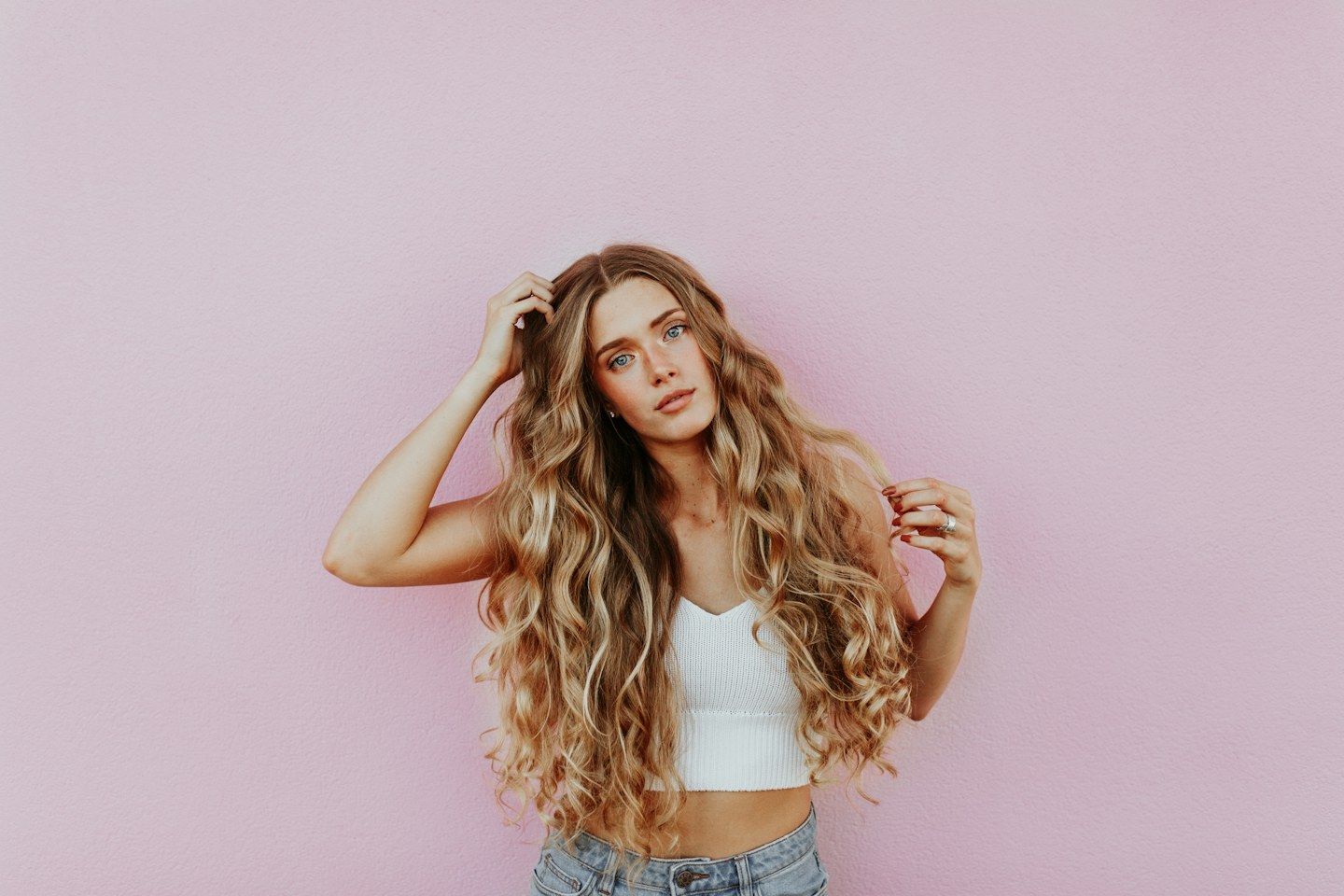Mulher de cabelo loiro dourado, longo e ondulado, olhando para a câmera diante de uma parede rosa